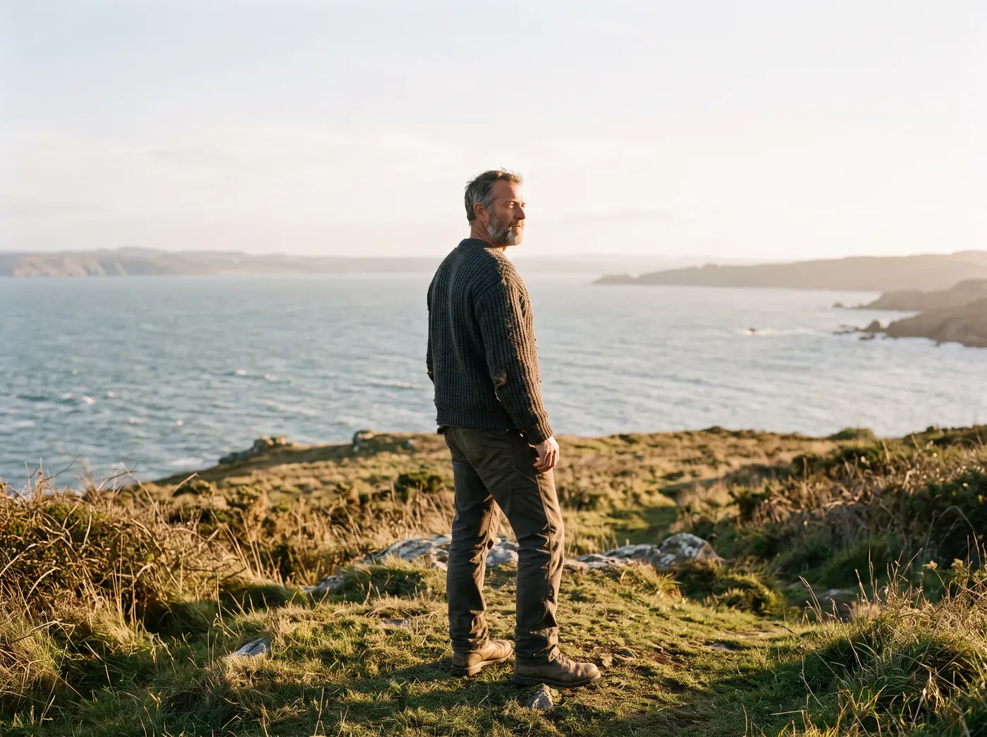 A man on a grassy headland, looking across a bay toward a shore he knows.